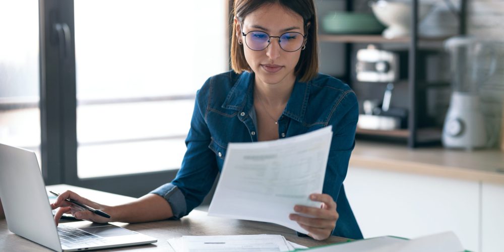 Pretty young business woman working with computer while consulting some documents in the kitchen at home.