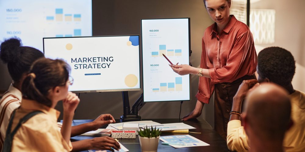 Portrait of young woman giving presentation on marketing trends to team in dimly lit office