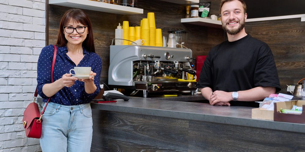 Young male worker talking to woman customer in coffee shop, near counter with cup of freshly prepared cappuccino coffee. Small business, food service occupation, staff, work concept